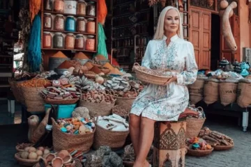 A woman in a white embroidered dress sits at a traditional Moroccan market surrounded by baskets filled with spices, herbs, and handmade goods, capturing the essence of a 4 days desert tour from Marrakech.