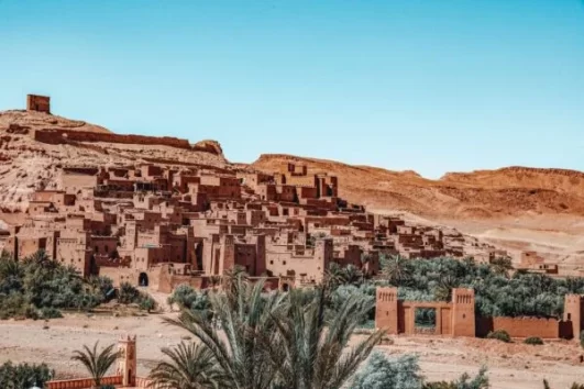 Historic Kasbah Ait Ben Haddou on the Chefchaouen to Marrakech route, featuring ancient clay buildings, desert landscapes, and palm trees under a blue sky.