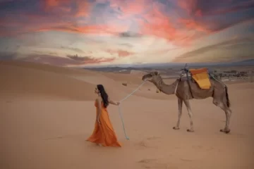 A traveler in an orange dress leading a camel through the golden sand dunes of the Sahara Desert during sunset, a highlight of the 9 Days Tour From Casablanca.