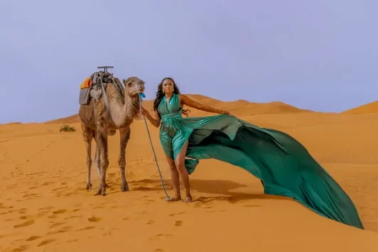 Elegant woman in a flowing green dress standing beside a camel in the golden Sahara Desert during the Morocco Chefchaouen to Marrakech​ tour