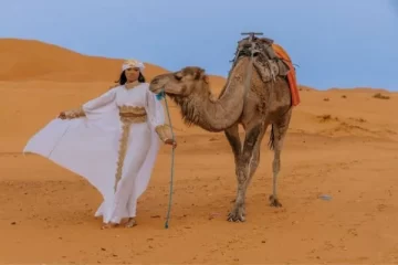 A woman in an elegant white outfit walking with a camel in the golden dunes of the Sahara Desert in Merzouga, capturing the essence of a Camel Trekking Sahara Tour Merzouga.