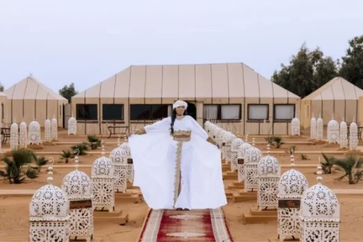 Luxury desert camp in Morocco with a woman in traditional white attire standing on a decorated pathway, surrounded by lanterns and beige tents in the Sahara. Errachidia to Marrakech desert experience.