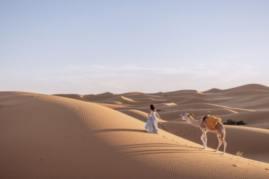 A woman in a flowing white dress walks with a camel across the golden sand dunes on the Errachidia to Fes desert tour in Morocco.